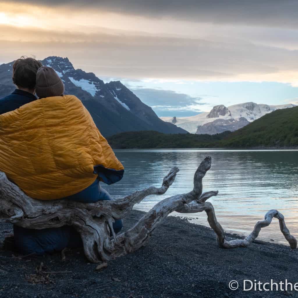 A couple keeping warm with a sleeping bag next to a cold lake and mountain view