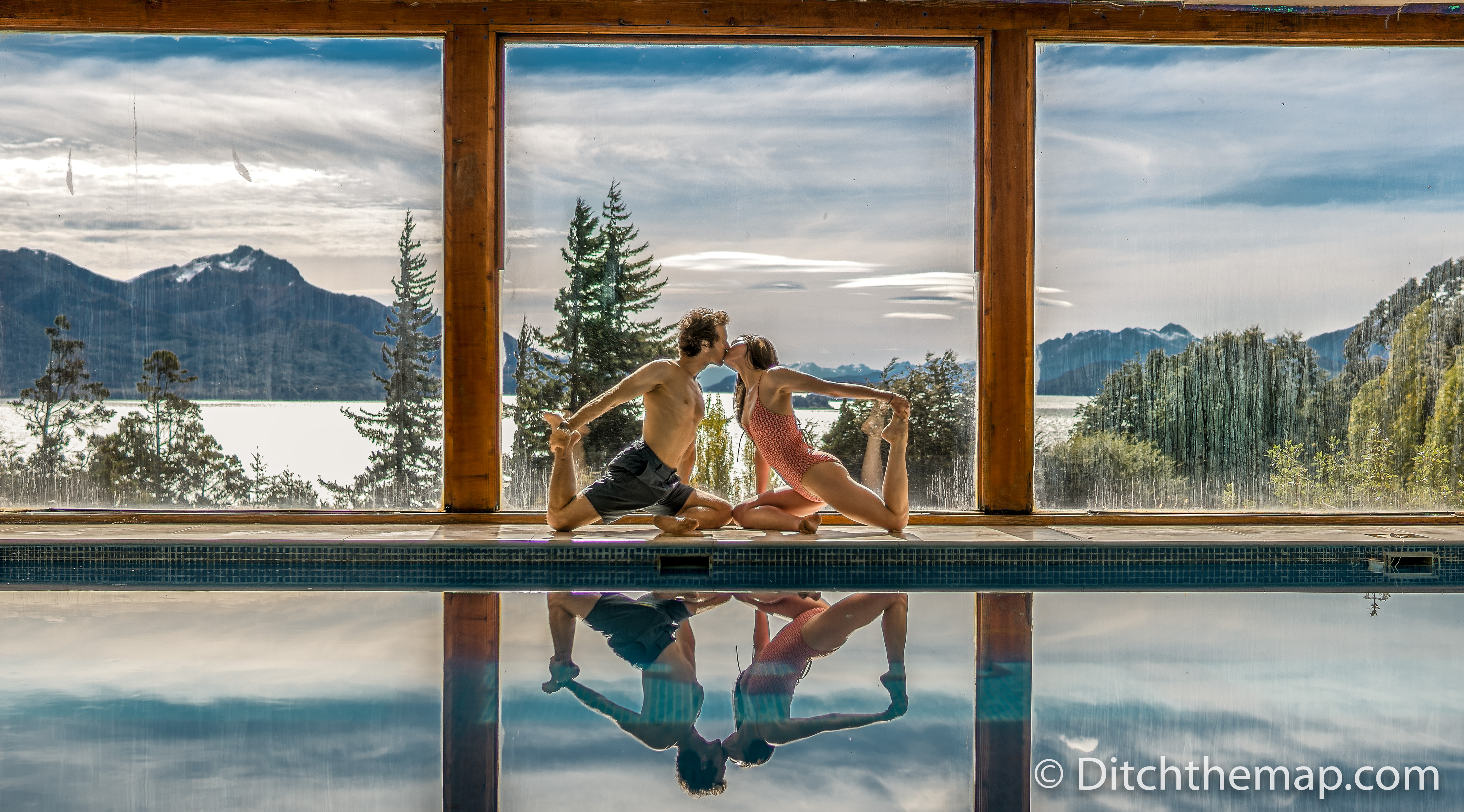 A man and a woman kissing while in a yoga pose next to a pool with a mountain landscape