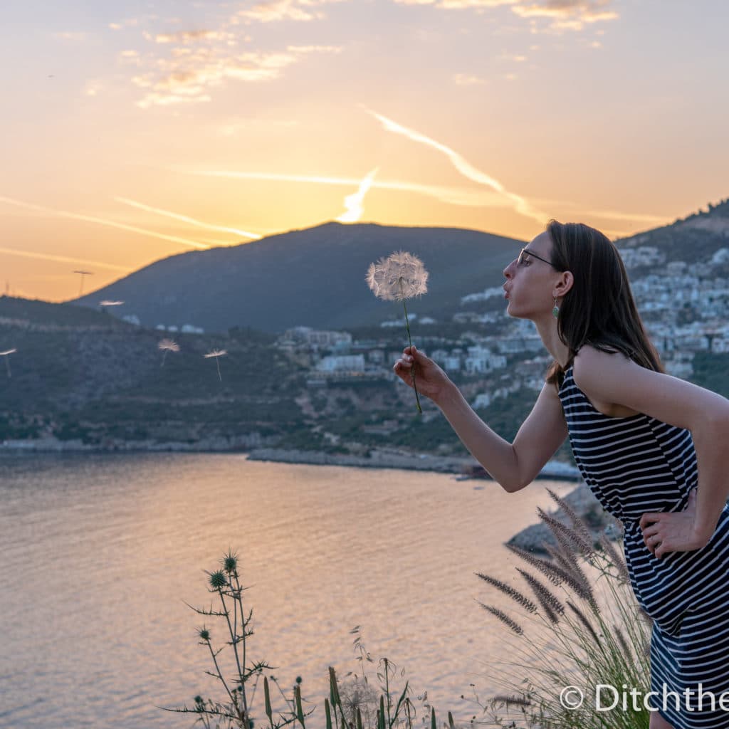 A girl blowing a dandelion during sunset next to a lake and mountains