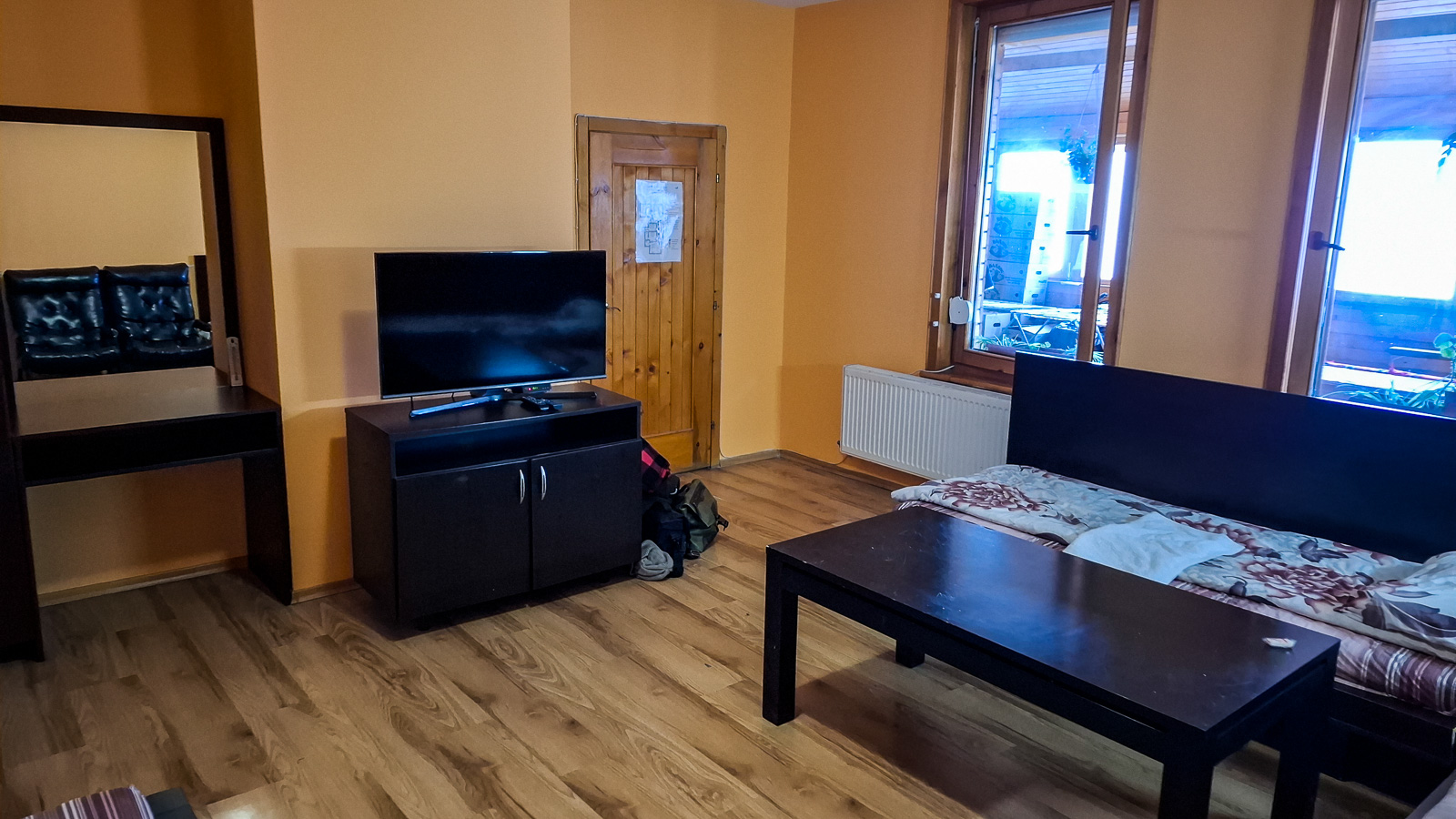 room interior with a shelf, tv, door, windows, table, a mirror and a bed